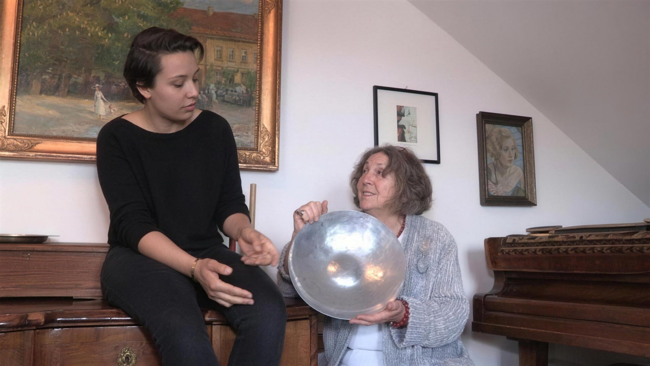 Filmszene: Eine junge Frau und eine ältere Dame sprechen über eine Silberschale, im Hintergrund Gemälde und ein Klavier