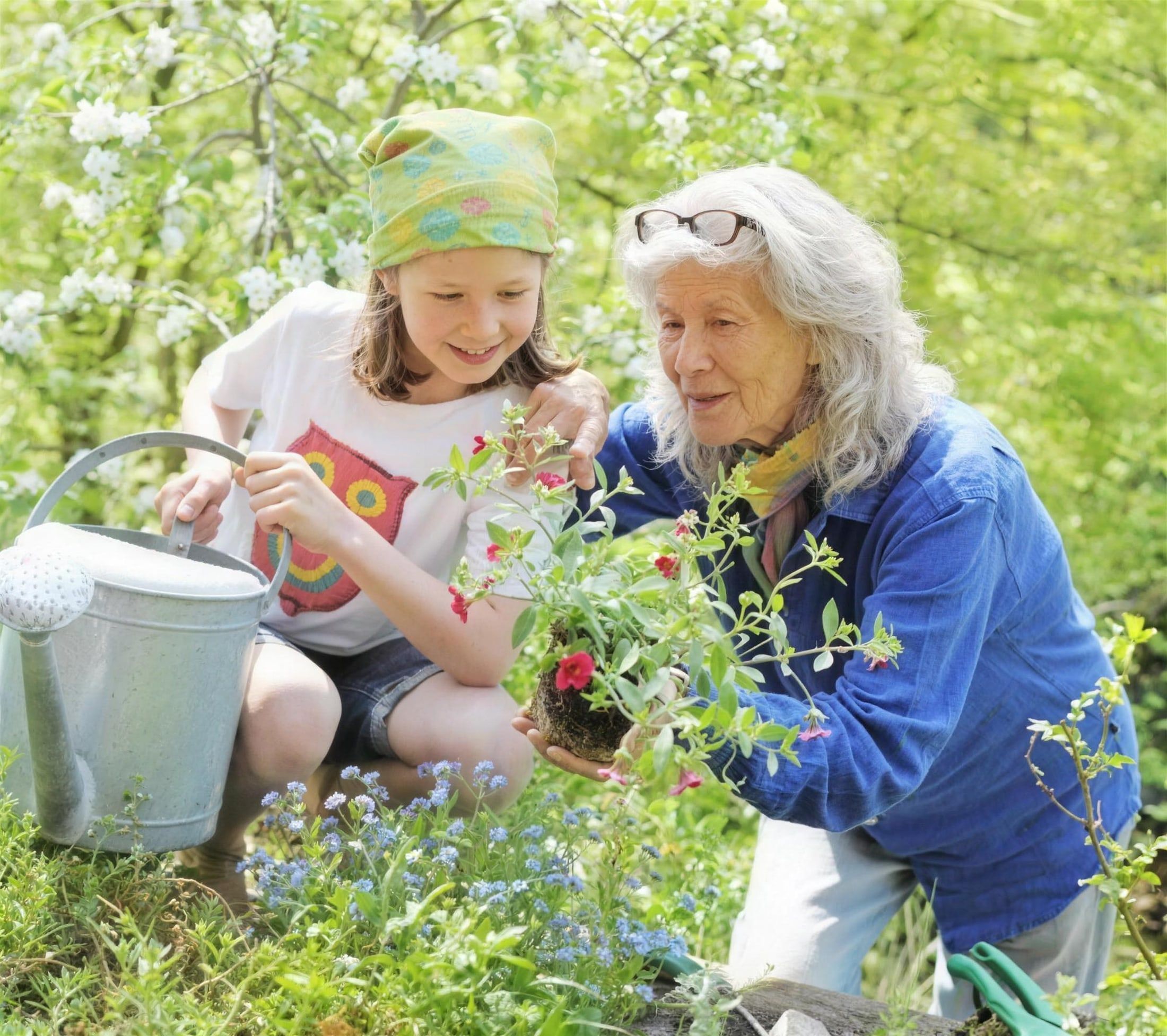Großmutter und Enkelin pflanzen gemeinsam Blumen im blühenden Garten, daneben eine Gießkanne