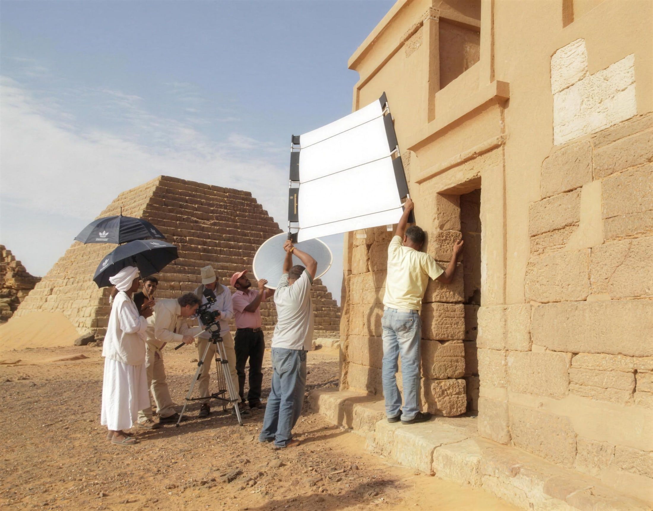 Filmteam mit Kamera, Reflektoren und Sonnenschirmen bei Dreharbeiten an einer antiken Pyramide in der Wüste
